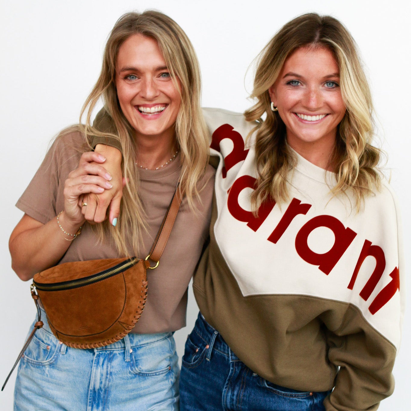 Two women posing together, one holding a brown leather bag, against a white background.